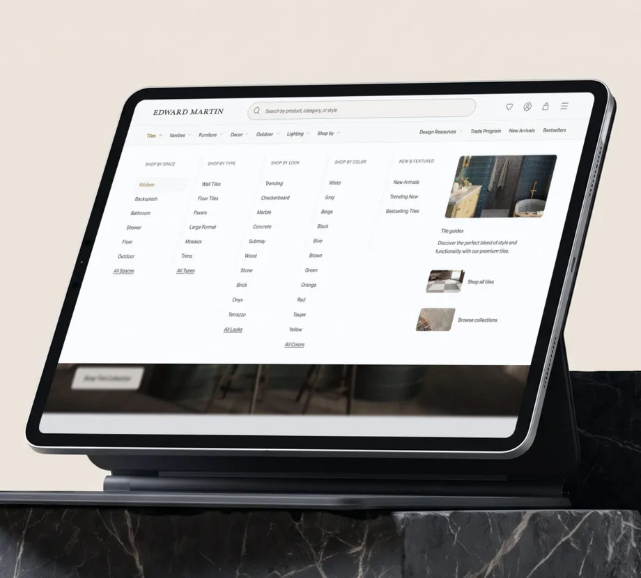 The Tiles megamenu open on an iPad in a keyboard case, standing on a black marble ledge — five columns of promoted routes, Shop By Space with Kitchen highlighted, Shop By Type, Shop By Look, Shop By Color and New & Featured, beside a Tile guides panel